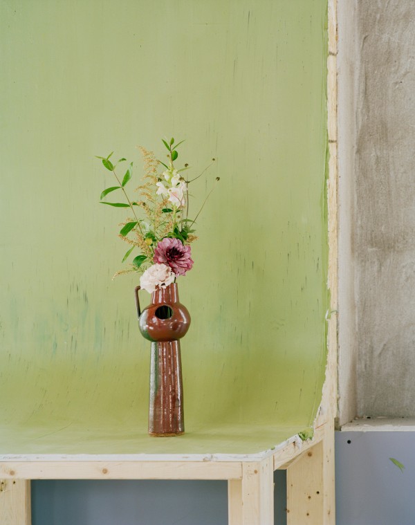 Waard Ward in collaboration with Nicolas Fleming & Darren Rigo, 'Flowers for Shirin Tawadros', 2021 / A photograph of a tall brown vase with greenery and a few white and pink flowers standing on a green surface which juts out from the wall.