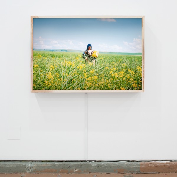 Kablusiak, Piliutiyara (Robin Hood), 2021. Image Description: Lightbox with a photograph of a female figure wearing lingerie in the middle of an open field of grass and flowers. It is a sunny day, and the blue sky reveals faint clouds in the distance. The figure stands with an ambivalent expression on their face and holds a large bag of Robin Hood brand flour.